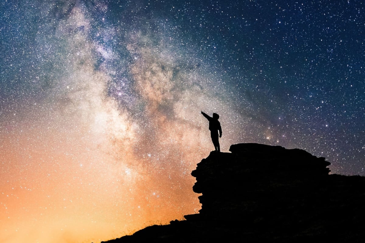 Lone person standing on top of rock, pointing with hand towards the Milky Way and the vast expanse of universe. Stunning silhouette of man gazing out into the cosmos. Power of dreams and aspirations.