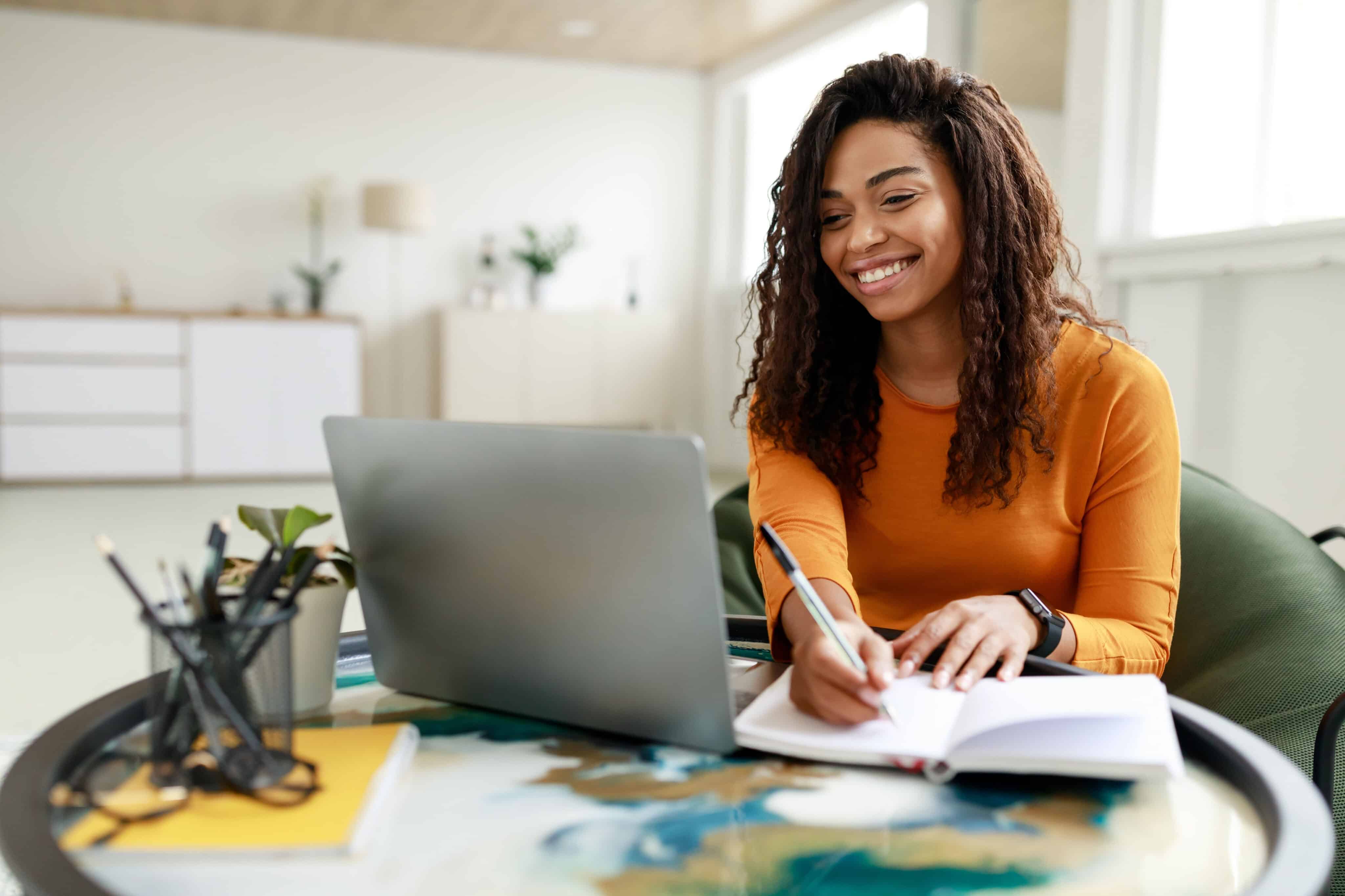 Smiling young African American woman sitting at desk working on laptop taking notes in notebook, happy millennial female studying online, watching webinar using computer and writing check list.
