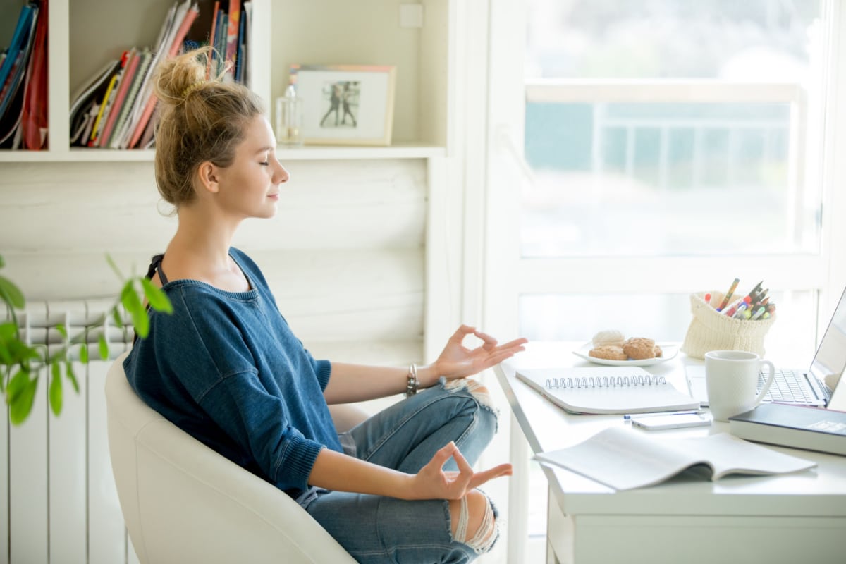 Portrait of an attractive woman in a chair at the table with cup and laptop, book, pencils, notebook on it. Lotus pose, concept photo