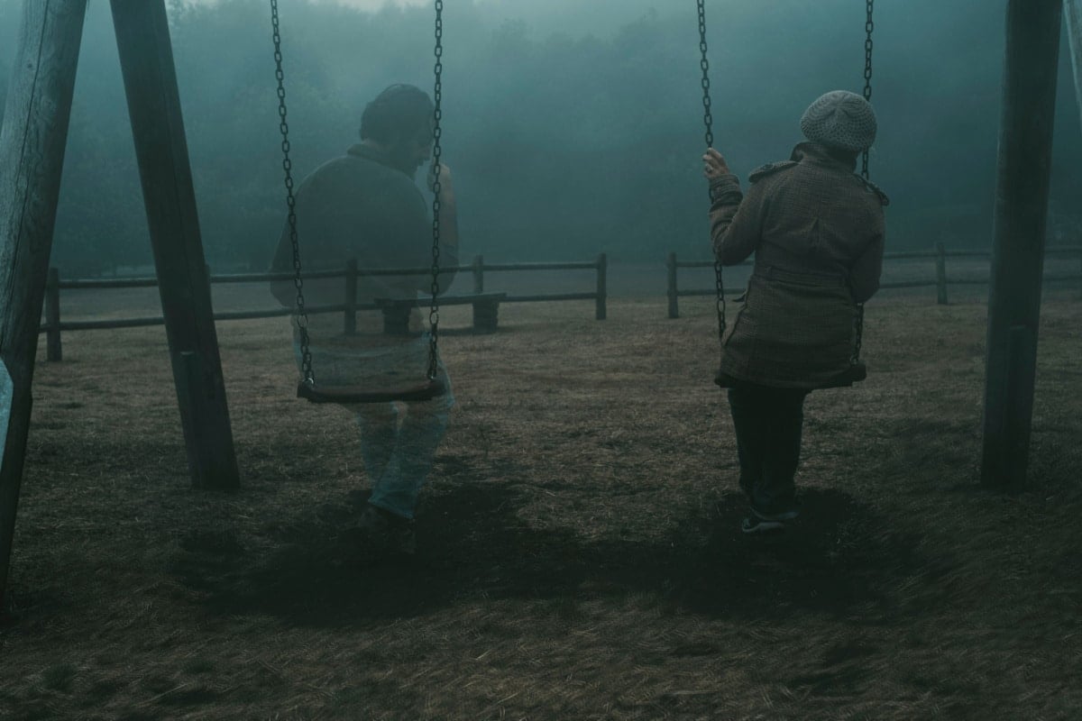 Concept of afterlife and memory for parents or lost love. Back view of woman sitting on a swing with ghost man near her outdoor in the park with fog. . Dead friend or husband concept. Life and death