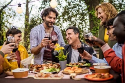 Cheerful diverse group celebrating with wine toast at alfresco dinner. Multicultural friends gathered around table with appetizers, enjoying outdoor meal in garden setting.