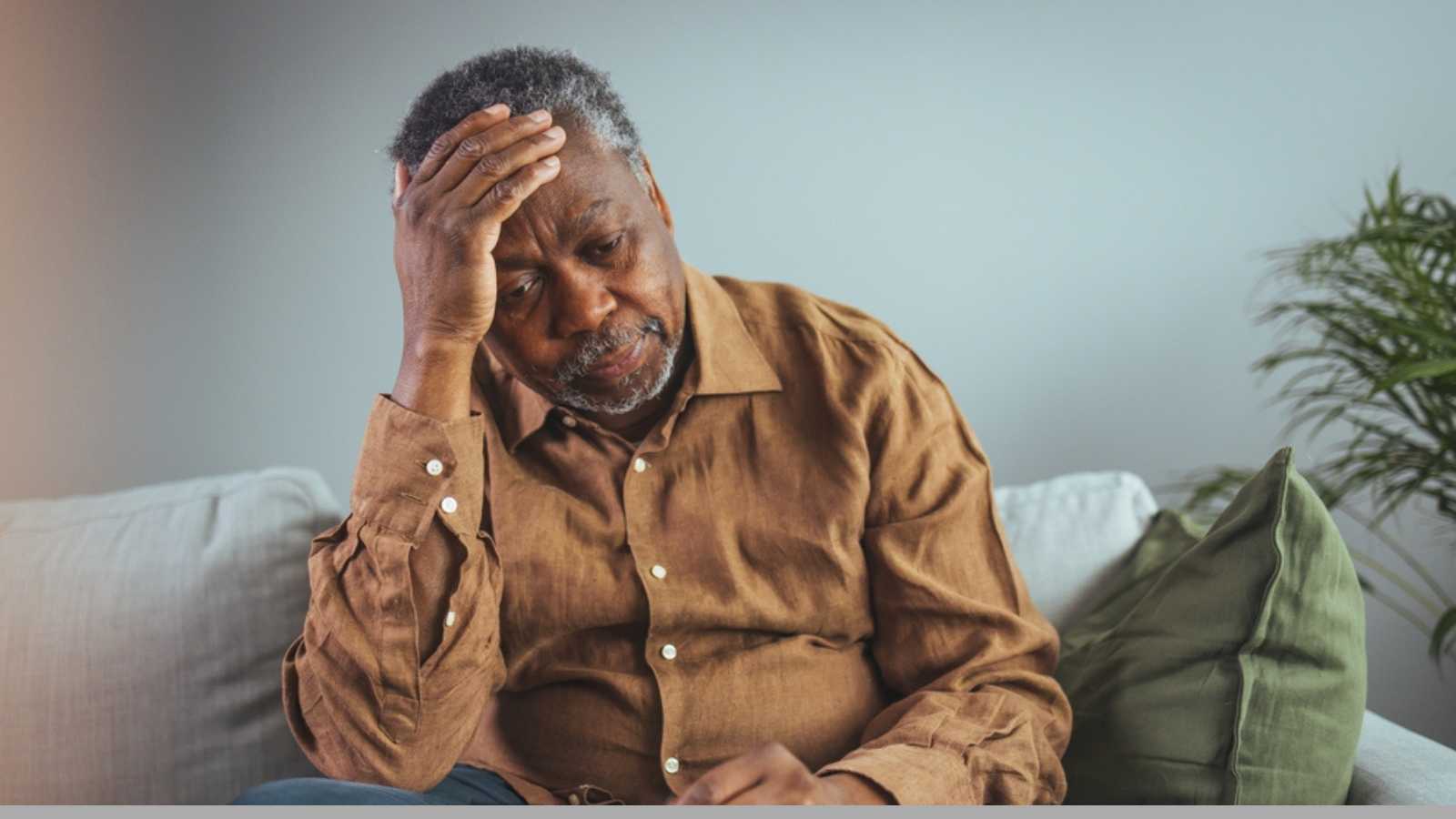 Elderly Black Man Experiencing Discomfort on Couch. Senior black male grips his glasses, showing signs of distress while seated alone on a sofa, reflecting potential health concerns in a home setting.