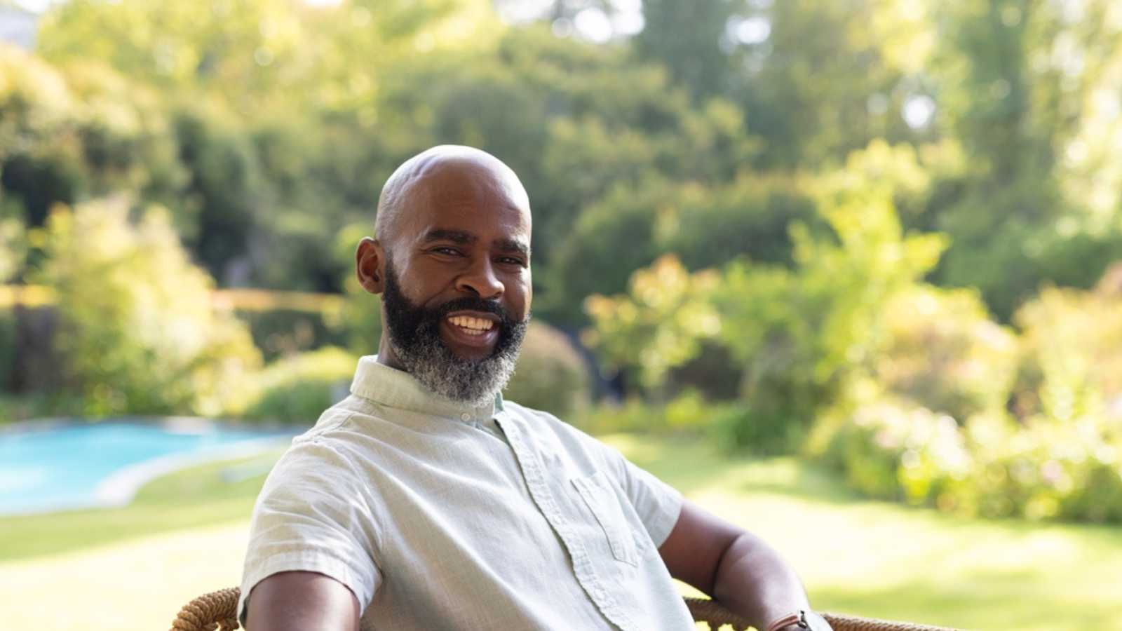 An African American senior man, wearing light clothes, sitting outdoors, smiling. Showcasing full beard and bald head, enjoying a sunny day with a relaxed posture, unaltered