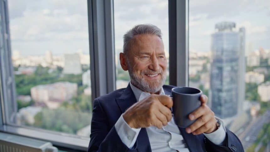 Wealthy executive holding coffee cup sitting by office center panoramic window closeup. Smiling aged ceo relaxing after successful workday. Happy elderly businessman enjoy beverage looking cityscape, senior man happy