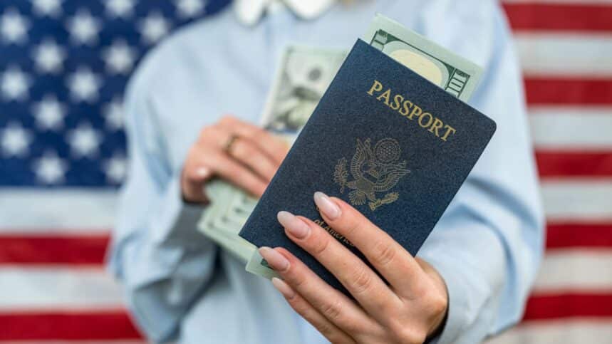 Beautiful woman in a classic shirt holds an American passport with money dollars on the background of the USA flag