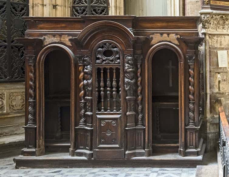 An antique wooden confessional booth with two entrances, and a crucifix inside one of the chambers.